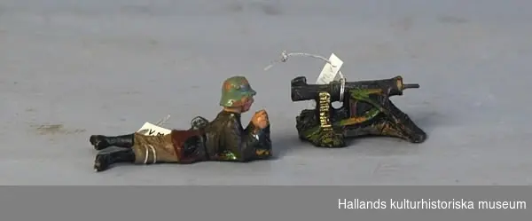 Leksakssoldat av trämassa på ståltrådsskelett i tysk uniform. Soldaten ligger ner och skjuter med en kulspruta. Målad i grönt, brunt, svart, silver och skärt.