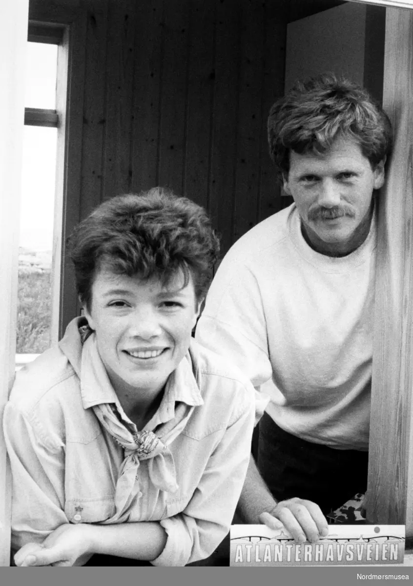 Karin Stokke og John Arne Selnak på jobb i bompengeluka ved Atlanterhavsveien mellom Averøy og Eide i Møre og Romsdal. En veistrekning på 8.274 meter. Anlegget består av åtte broer på til sammen 891 meter og går over holmer og skjær. Veien kostet 122 millioner i 1989 og var delvis bompengefinansiert. Anlegget ble åpnet 7. juli 1989 og bommen ble fjernet i juni 1999. Atlanterhavsveien er landets åttende best besøkte naturbaserte turistattraksjon (2004) med 283.500 besøkende, og veistrekningen er foreslått vernet i "Nasjonal verneplan for veger, bruer og vegrelaterte kulturminner." (Kilde: Wikipedia Norge). Bildet er fra avisa Tidens Krav sitt arkiv i tidsrommet 1970-1994. Nå i Nordmøre museums fotosamling.