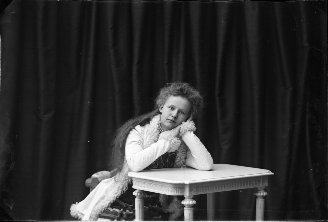Porträtt av en ung kvinna med långt hår. Hon sitter lutad över ett bord, vilar huvudet i händerna. Hon har en pälsbrämad ljus kappa på sig. Hennes namn är Mimmi Allard.