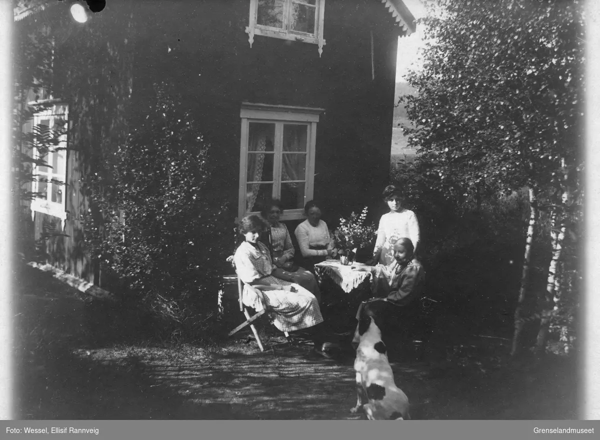 En gruppe kvinner fotografert sittende ved et bord ute i hagen, en hund i forgrunnen.