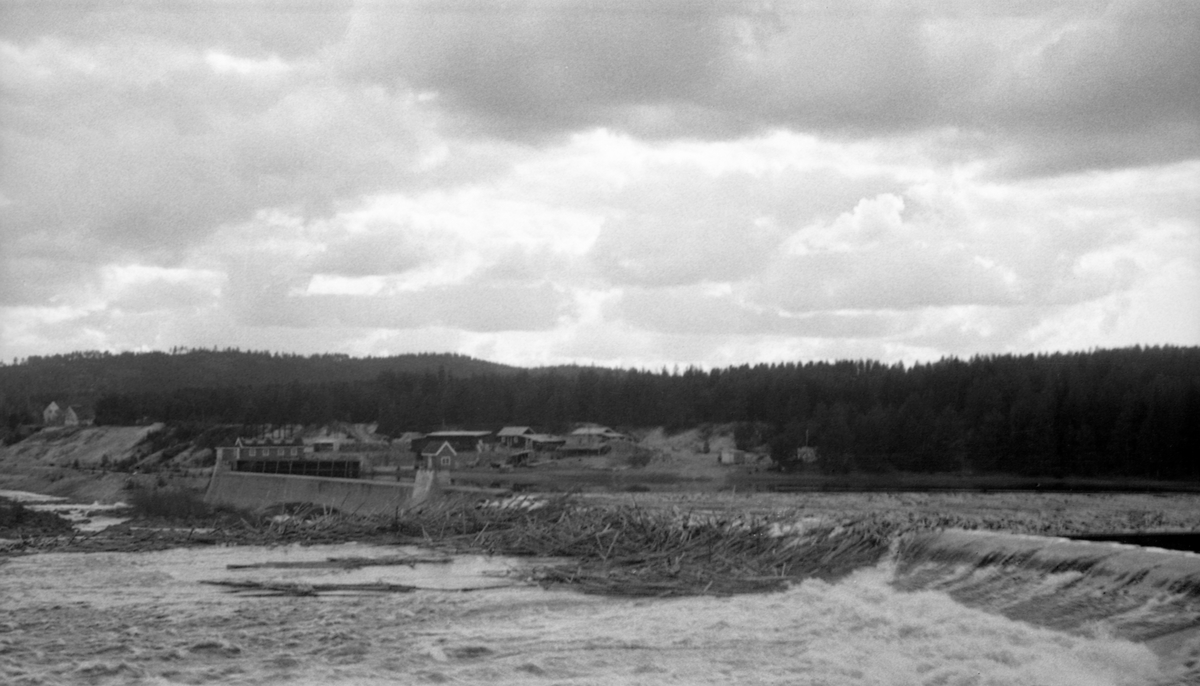Skjefstadfossen i Glomma, nærmere bestemt i Heradsbygda i Elverum, Hedmark, fotografert i begynnelsen av mai 1944, på et tidspunkt da det var forholdsvis lite vann i elveløpet. Øverst i denne fossen hadde man i 1909-1910 bygd et elvekraftverk med en terskeldam som skulle stuve opp vann til turbinrøret. Overløpsdammen var 312 meter lang, utført i betong og støpt slik at damkrona var noe lavere i den østre enn i den vestre enden. Hensikten var å skape et «sug» i overflata som trakk fløtingstømmeret mot den østre delen av elveløpet. Av samme årsak brukte man å legge ei lense, et flytende stengsel lagd av sammenkjedete tømmerstokker, som skulle hindre at fløtingsvirket drev inn i inntaksbassenget for vann til kraftstasjonen, som lå i den vestre delen av elveløpet. Da dette fotografiet ble tatt, i slutten av april 1946, hadde det samlet seg en del tømmer langs den nedre delen av ledelensa på dammen, og noe av tømmeret derfra hadde lagt seg i nedfallssona under overløpsdammen. Til venstre i bildet ser vi den nevnte inntaksdammen med påstående lukehus.