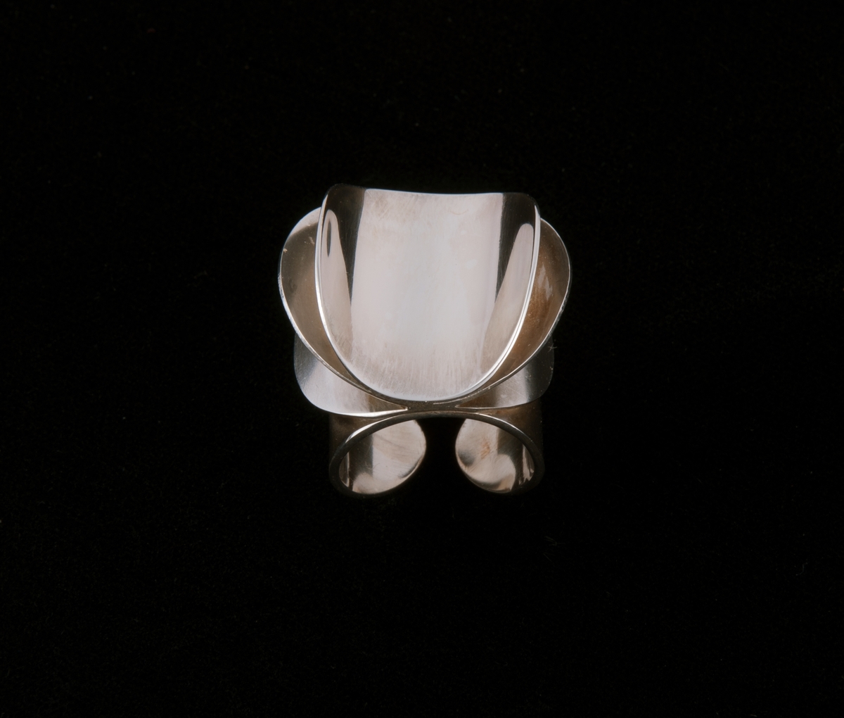 Består av tre like store sirkulære sølvringer som er satt sammen langs en midtakse. På hver siden av aksen er ringene bøyd oppover i ulik grad, symmetrisk på hver side. Dekoren er festet på en fjerde rund form som danner ringen rundt fingeren.