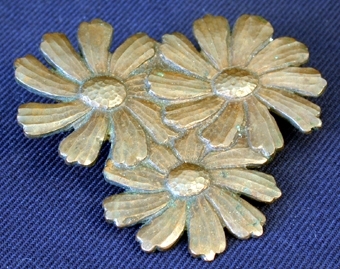 Platt kopparstycke format till tre stycken prästkragsliknande blommor som går in i varandra och tillsammans bildar en triangelliknande form. På broschens baksida sitter en nål med säkerhetsnålsfunktion, med vilken man fäster fast broschen på hatten.