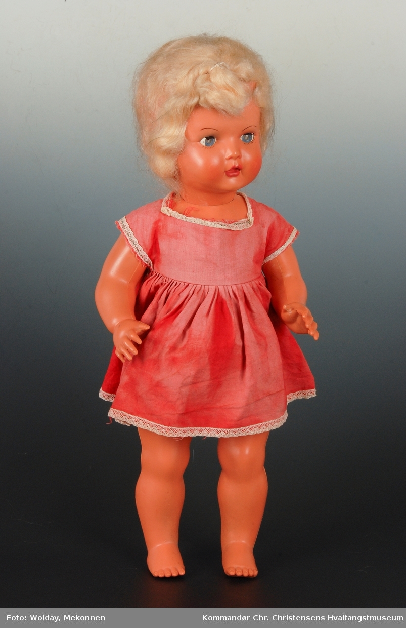 Dukken har rød kjole med hvit blondkant nederst. Blondt, syntetisk hår.