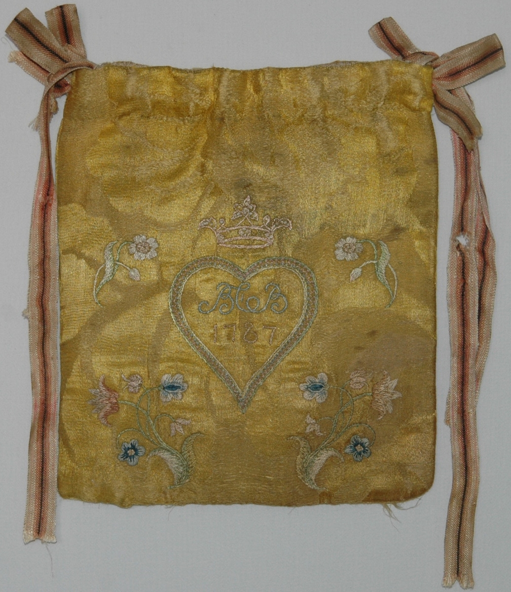 Handväska av gult sidendamasttyg med broderi i flera färer. Märkt på ena sidan BCB 1787 i ett hjärta under en grevlig krona. På andra sidan en broderad blomma. Fodrad med linnetyg.