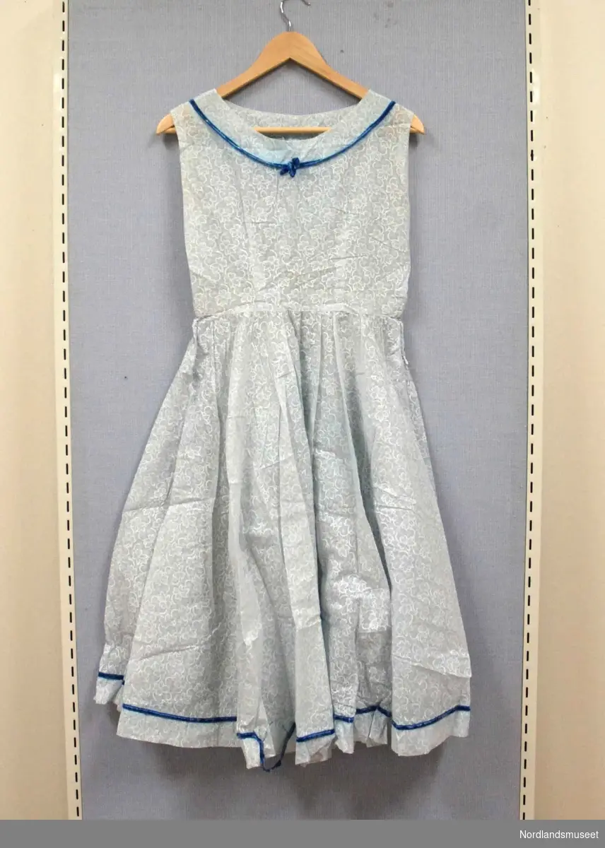 Lys blå kjole i tynt, delvis gjennomsiktig stoff. Hvitt blomstermønster. Blått fløyelsbånd i øverst på brystet og nederst på skjørtet. Glidelås i ene siden.