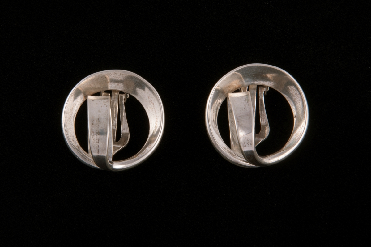 Blankpolerte øreklips av flathamret sølvbånd av varierende bredde. Bøyd i sirkel og ført ned i en løkke inne i sirkelen der endene er loddet sammen.