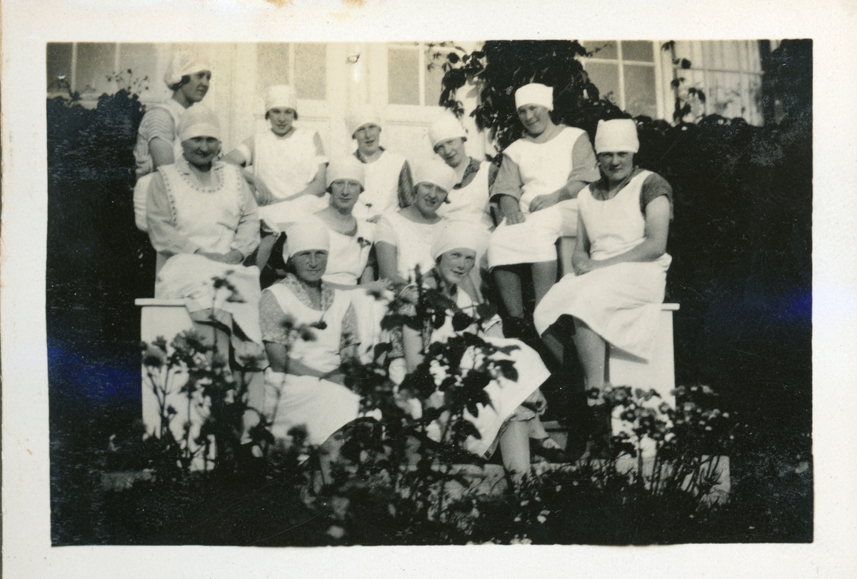 Elever fra husmorskolen på Skansgården, Kongsvinger, 1928. Elleve unge kvinner i forklær og tørklær sittende i trapp foran huset.