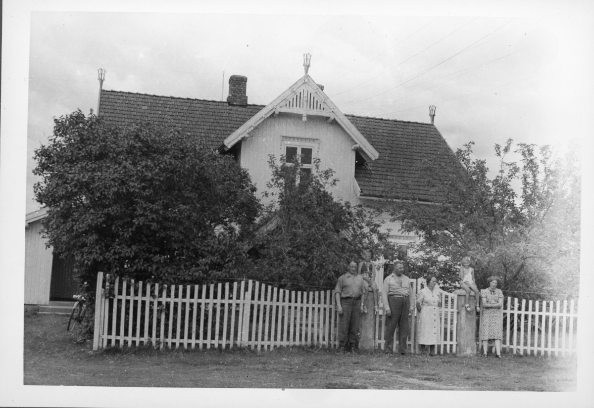 Familie med barn og voksne foran port stakittgjerde og sveitserstilshus med hage. Trolig 1960-tall.