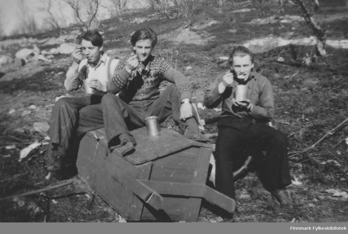 Tre menn sitter på en trekasse ute i naturen og spiser suppe. Området er antakelig i Børselv og tidspunkt på 1940-tallet.