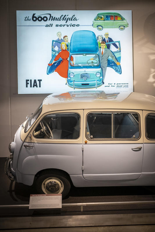 En Fiat stilt ut på Norsk kjøretøyhistorisk museum, bak bilen er en reklameplakat for bilen.