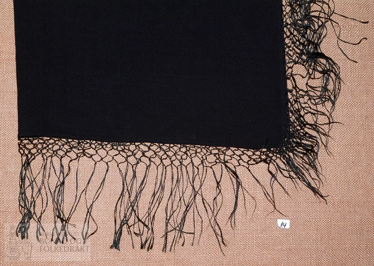 Svart ullmusselin med svarte silkefrynser, 12,5 cm lange med fire knyterader. Falda med maskin i to sider, jarekant i dei to andre sidene.  Storleik:  82 x 80 cm.