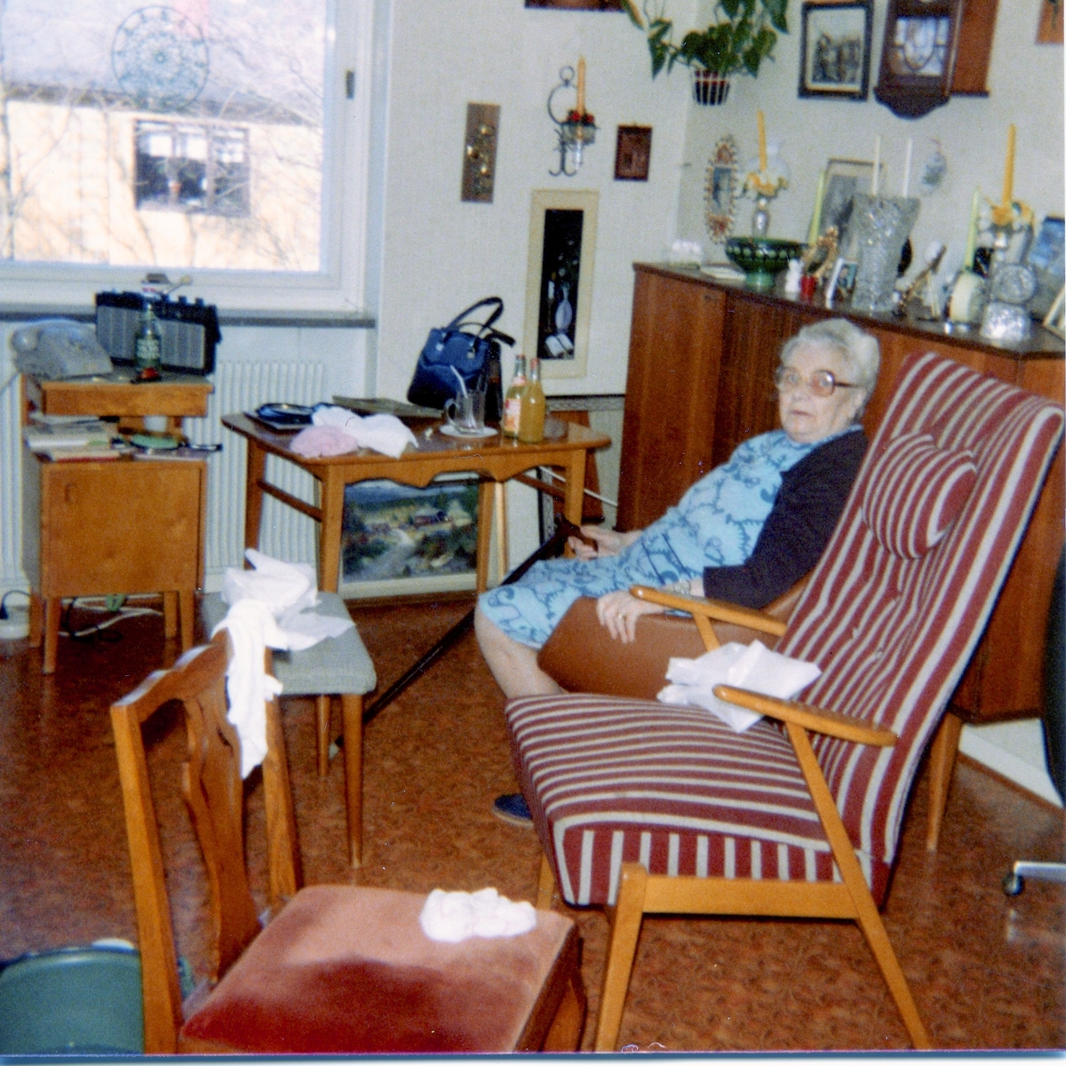 En äldre kvinnar sitter i en fåtölj i sitt pensionärsrum på Brattåshemmet 1979. Kvinnan håller en käpp i handen. Troligtvis är möblerna hennes egna.