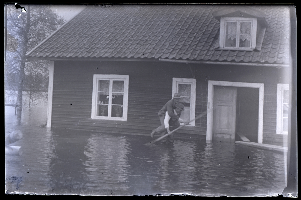 Översvämning.
Ett bostadshus står till viss del under vatten.
Två män utanför, varav den ene bär den andre på ryggen.