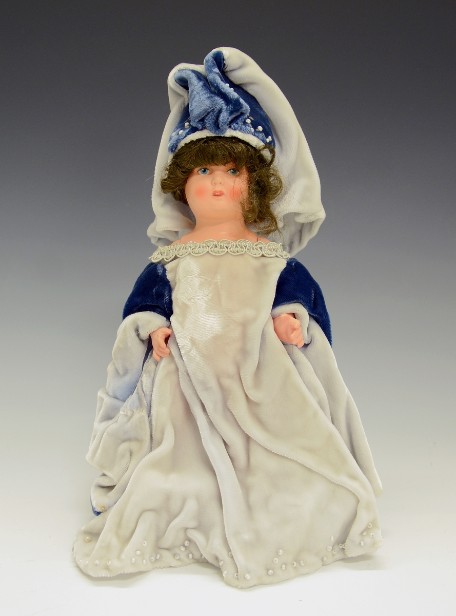 Dukke av plast i blå kjole. Dokka skal forestille gotisk middelalder ca. 1200-1480 og den sitter på en hvit hest med rødt seletøy.