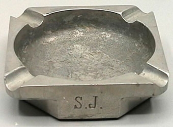 Fyrkantig askkopp av grå metall med fördjupningar för att lägga cigaretten i varje hörn.
S.J instansat på långsidorna.
Askkoppen har sexkantig fot.