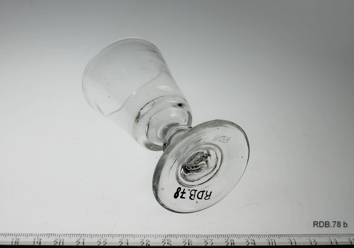 Drammeglass med "knapp" på stetten. Glasset er videre øverst enn nederst. Et merke under bunnen viser at glasset er handblåst.