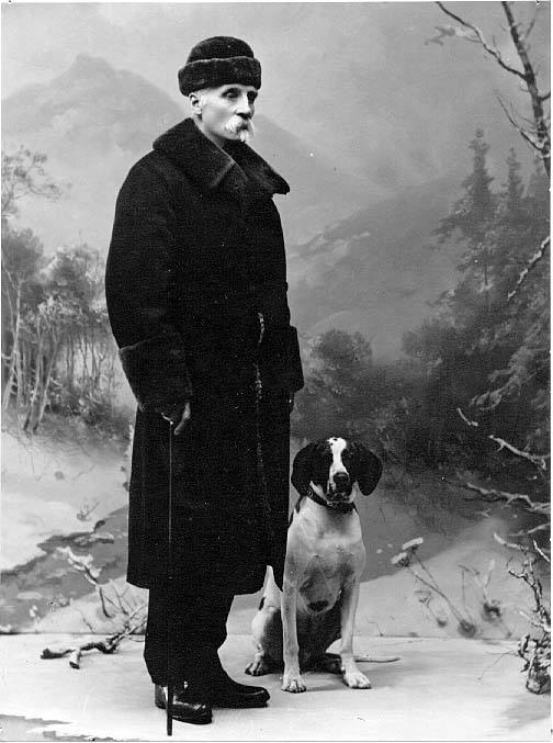 Porträtt av vinterklädd man i helfigur. Pälsmössa på huvudet och en käpp i höger hand. En hund sitter intill honom. I bakgrund en fond med vintrigt fjällandskap.