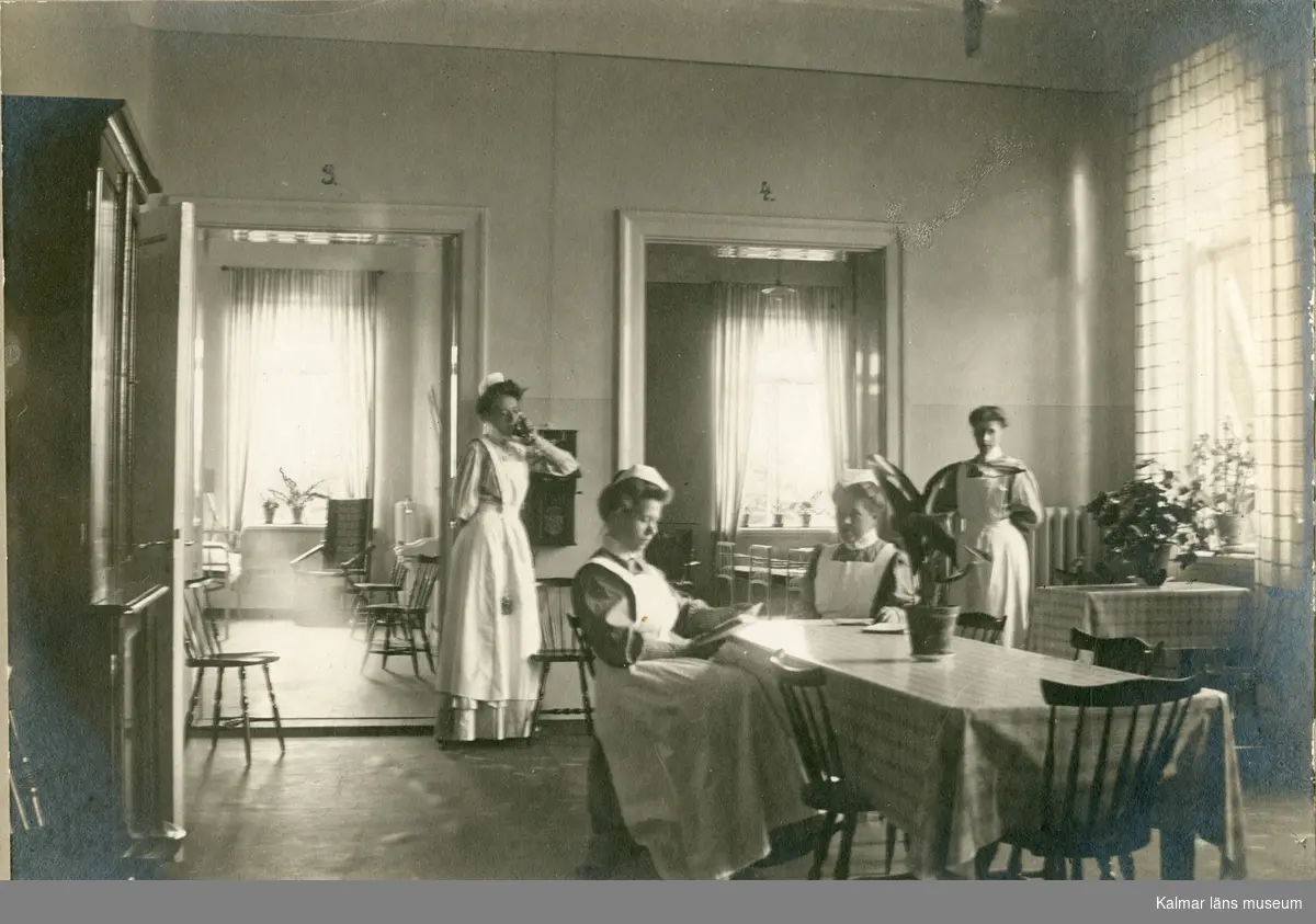 Motivet föreställer interiör från dagrum på Epidemisjukhuset på Lindö i Kalmar. Fyra stycken kvinnor i personalen uppehåller sig i rummet.