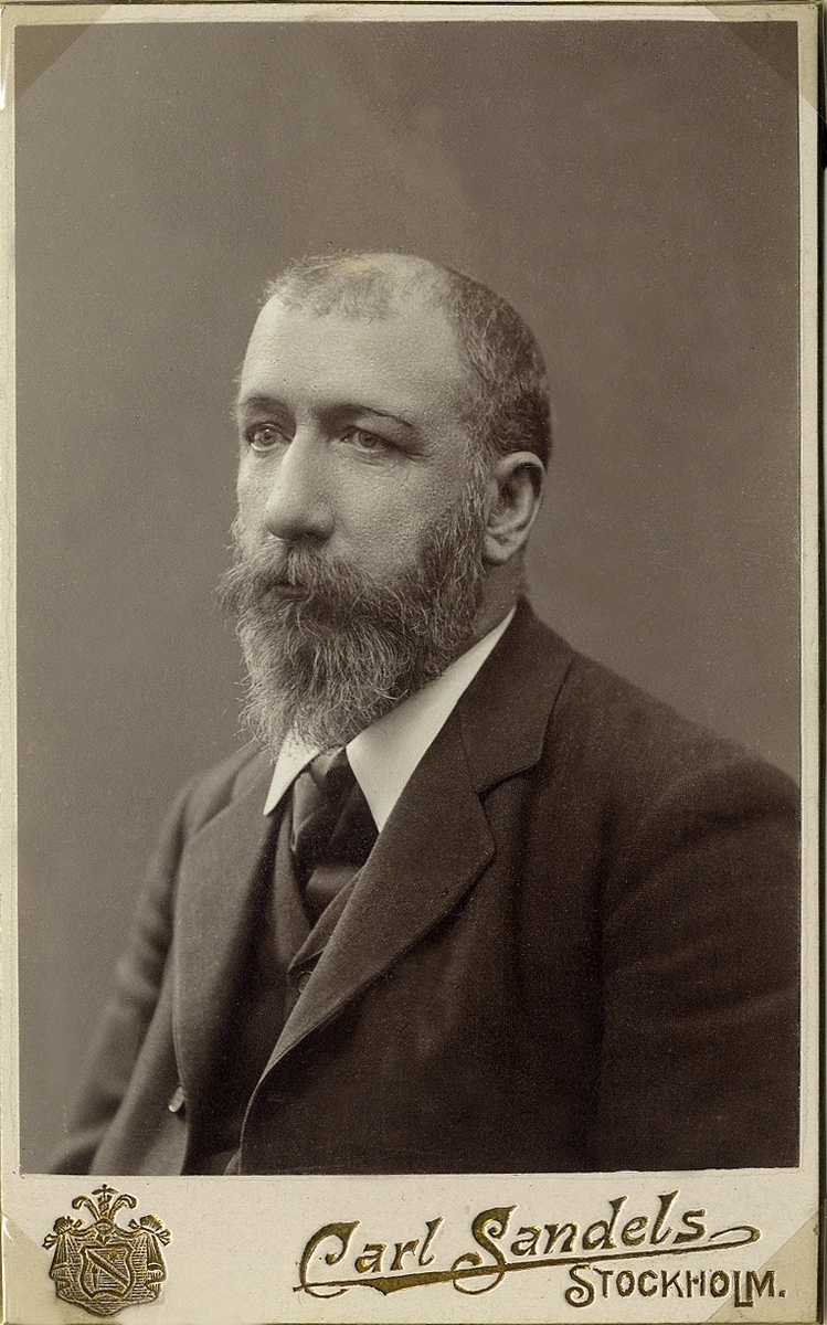 Porträttfoto av en skäggig man i mörk kavajkostym med väst och slips. 
Bröstbild, halvprofil. Ateljéfoto.