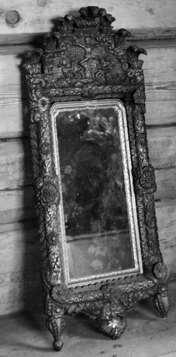 Speilramme av tre. Over speilet er det motiv av Jesus på korset, og over dette partiet går det ut seks palmetter. Ved bunnen går det ut to knotter på sidene.