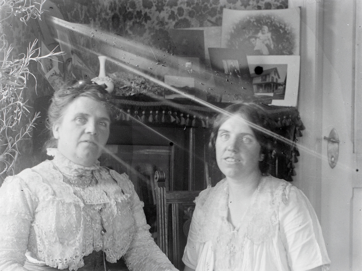 Portrett av to kvinner fra familien Aston i ei stue. I bakgrunnen står et skap med masse bilder på.