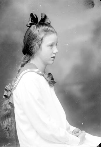 Porträtt av okänd sittande flicka i profil. Håret är flätat med rosett och en rosett på huvudet. Hon bär en ljus klänning med sjömanskrage.