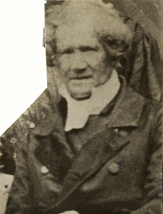 Foto av en äldre man med polisonger, klädd i rock, prästkrage m.m. 
Bröstbild, halvprofil, Ateljéfoto.