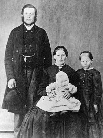 Gruppbild med familjen Gustav Johansson. Mannen står iklädd rock och väst med klockkedja. Han håller sin hatt i höger hand. Intill sitter hustrun med ett spädbarn i knät. En liten flicka i randig klänning står intill.