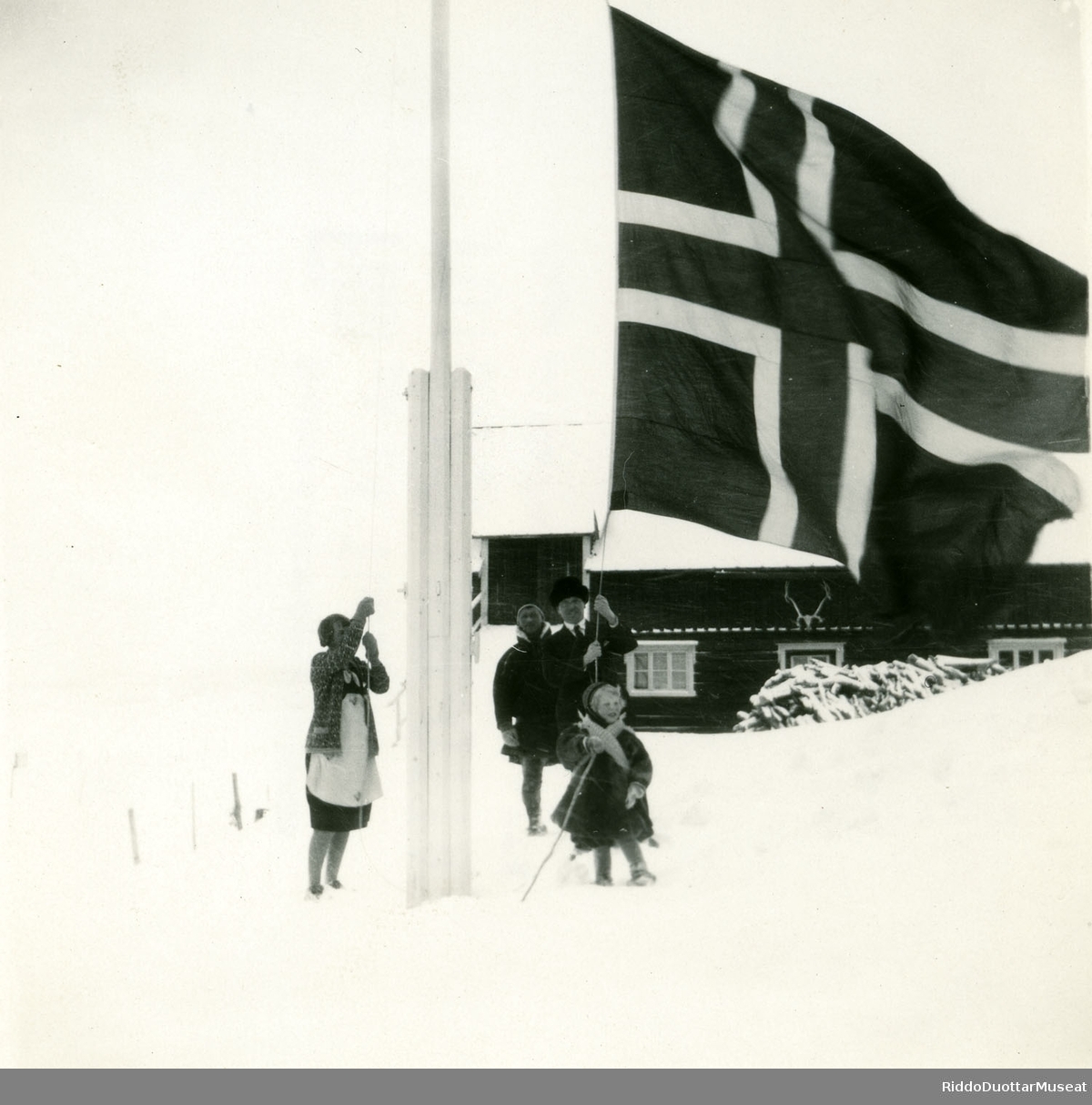 Njeallje olbmo norgga leavgga geassimen leavgastolpui.
Tre personer og en unge heiser opp det Norske flag.