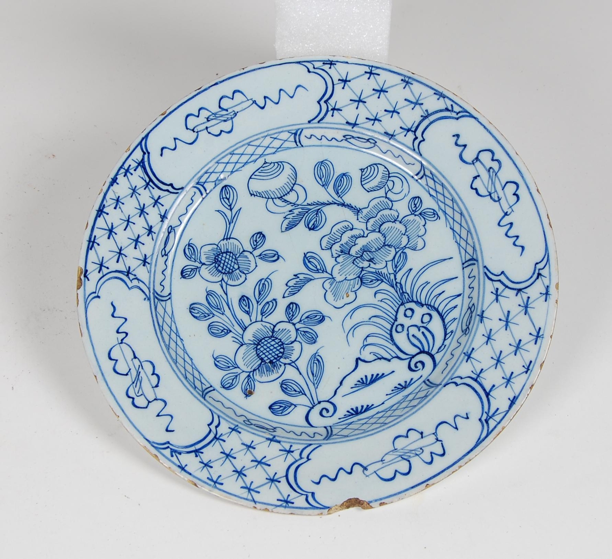 Hvitglasert tallerken med blå håndmalte blomstermotiv. Langs siden innvendig malt rutedekor.