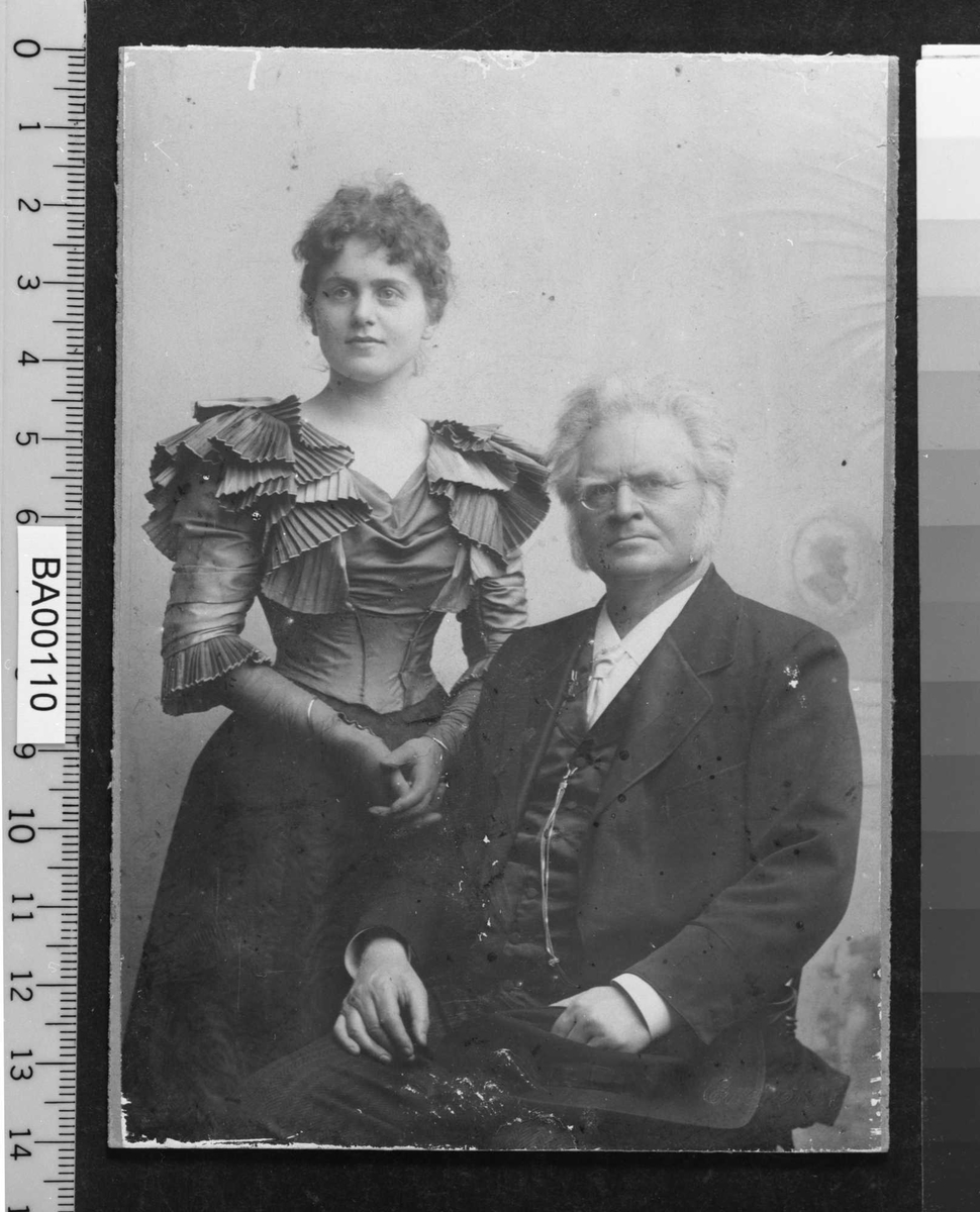 Atelierfotografi; dobbeltportrett, en ung dame stående i flott festkjole med hansker på, en eldre mann sittende på stol snudd 45¤ mot venstre inn foran henne. Han ser direkte på betrakteren.