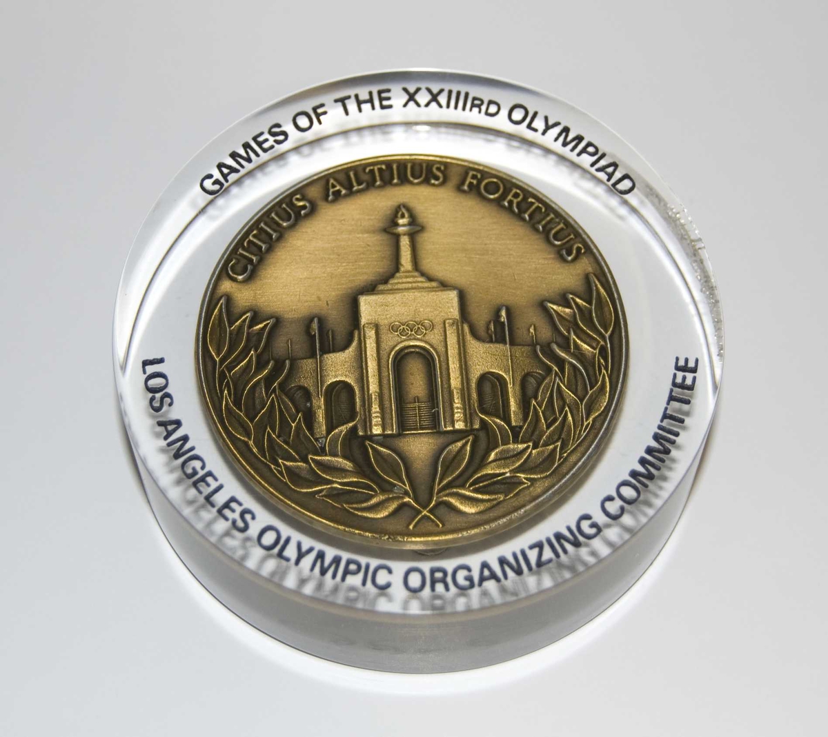Gullfarget medalje med emblemet for de olympiske sommerleker i Los Angeles i 1984 og motiv av det olympiske stadion i Los Angeles. Medaljen er innstøpt i pleksiglass.
