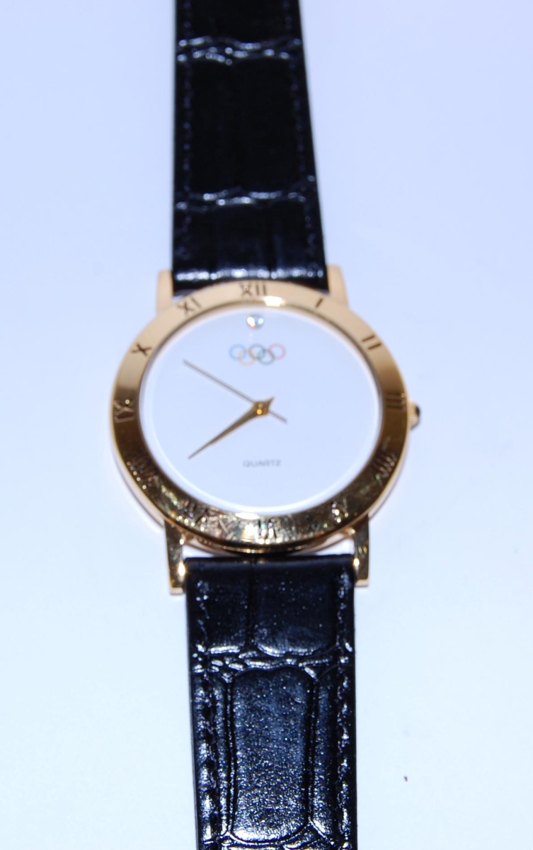 Gull- og hvitfarget armbåndsur med sort reim. På urskiven er det motiv av de olympiske ringene. Uret ligger i en svart, smal konvolutt, med olympiske ringer og tekst i gullfarge.