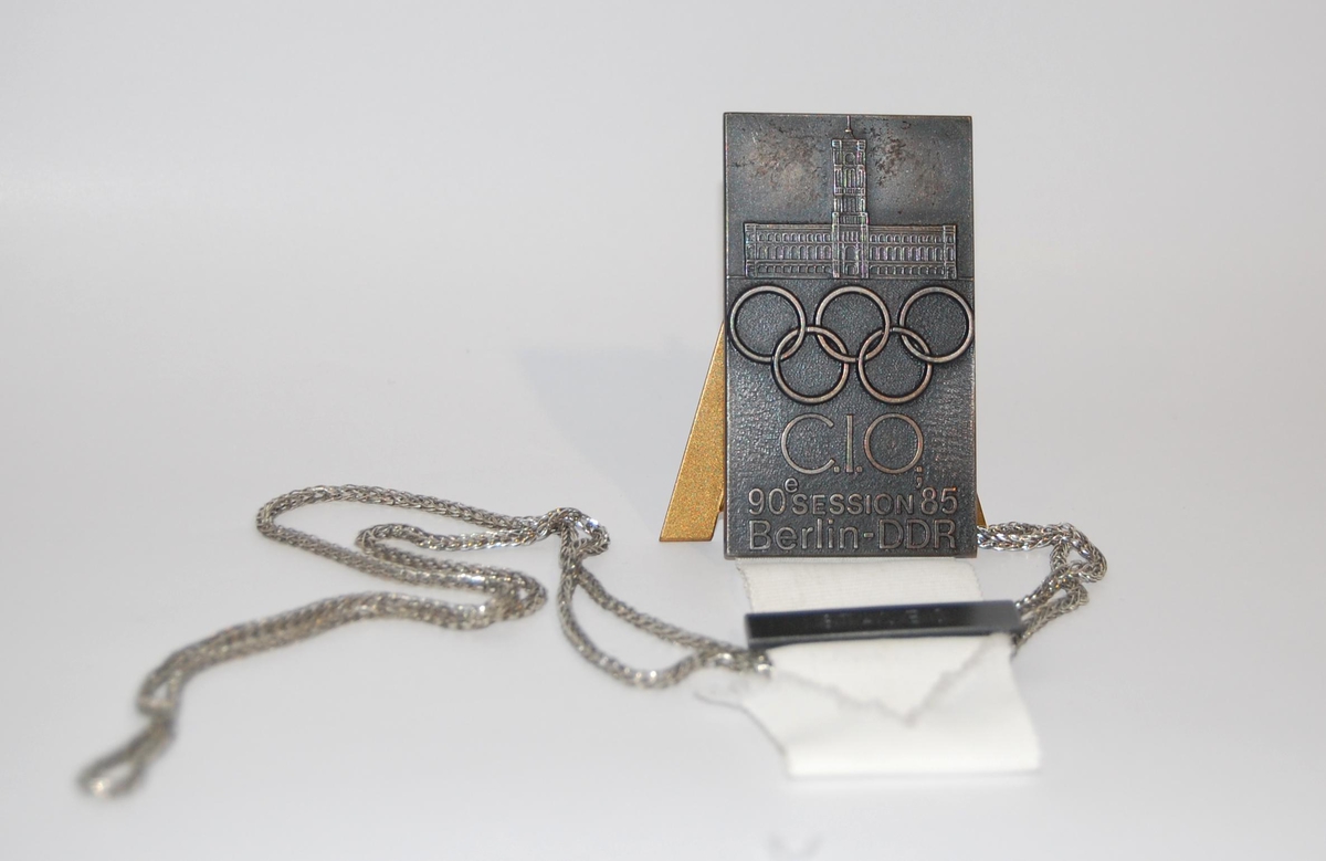 Jakkemerke med motiv av en bygning og de olympiske ringene. Til merket er det festet et hvitt bånd og en lenke av metall.