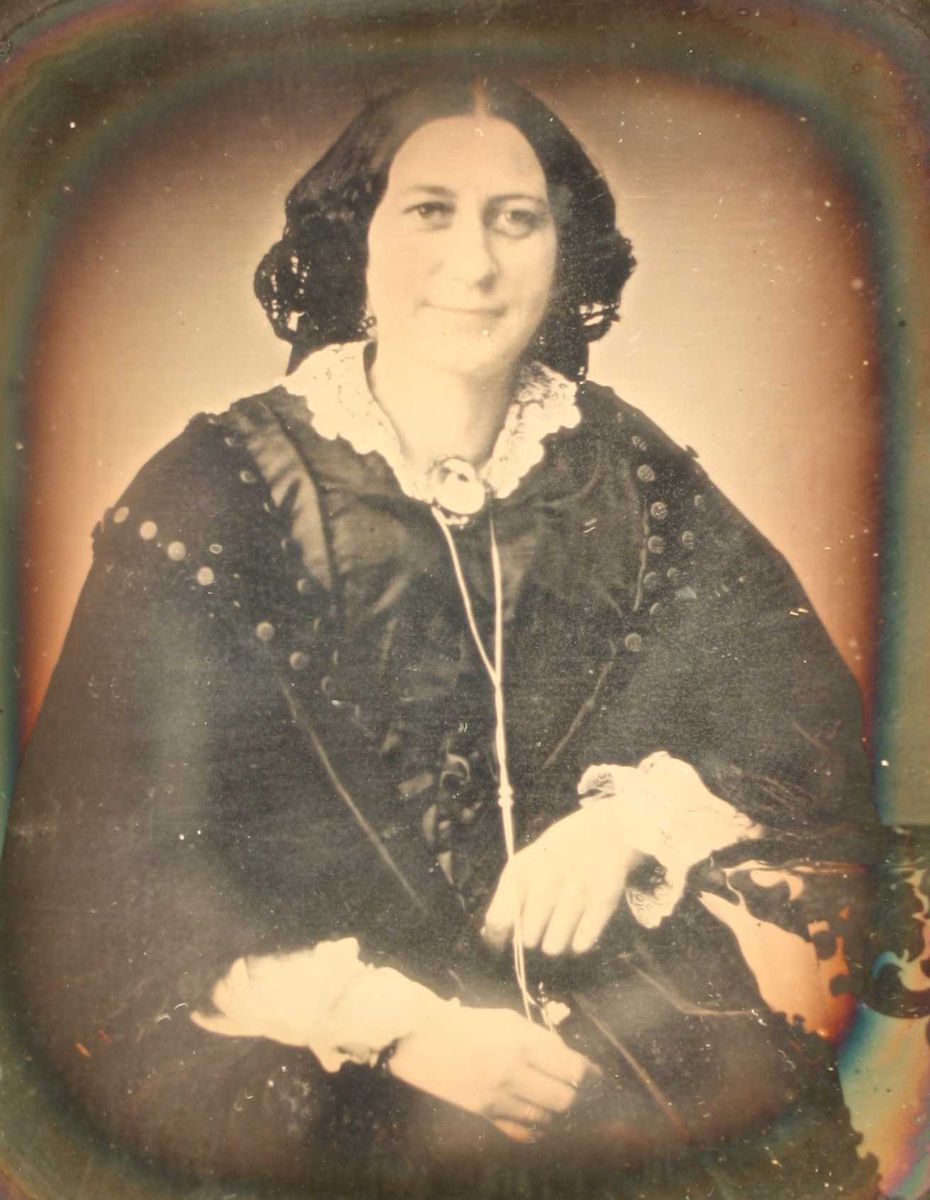 Portrett av Kaja Westergaard, 1856. Portrett av kvinne i en face, sittende, avsk. ved knærne, langt sort hår  med midtskill. Hvit blondekrave med brosje, holder med begge hender om en lang  lenke. Sort omramn. med gull inneramme.