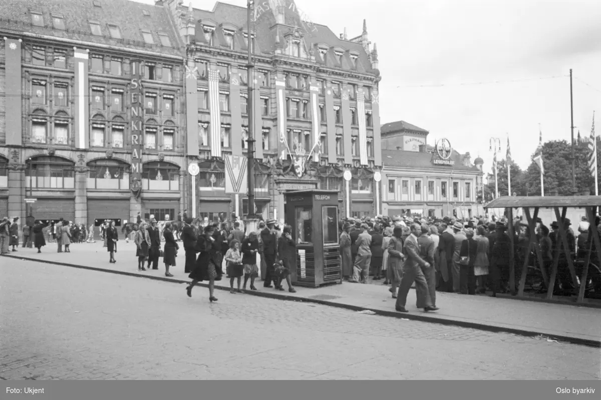 Folkeansamlinger på et pyntet Stortorvet 7. juni 1945 i anledning Kongens hjemkomst. Et pyntet GlasMagasinet,allierte bannere og flagg. Telefonkiosk.