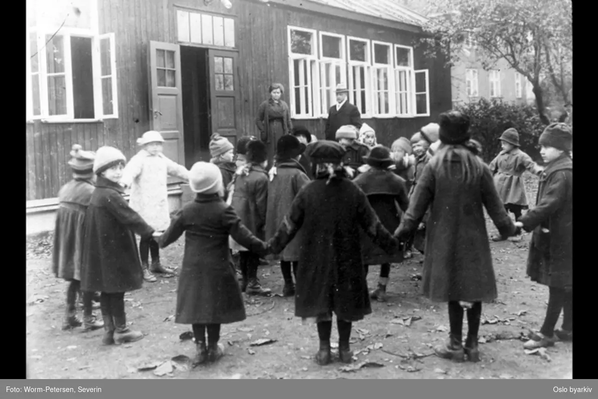 Sandaker friluftsskole. Barn som leker foran skolebygningen. I bakgrunnen Carl Schiøtz og skolens bestyrer Helga Kjerulf.