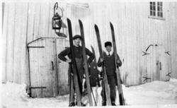 2 menn med hoppski foran uthus. Stor fjøslykt på veggen over