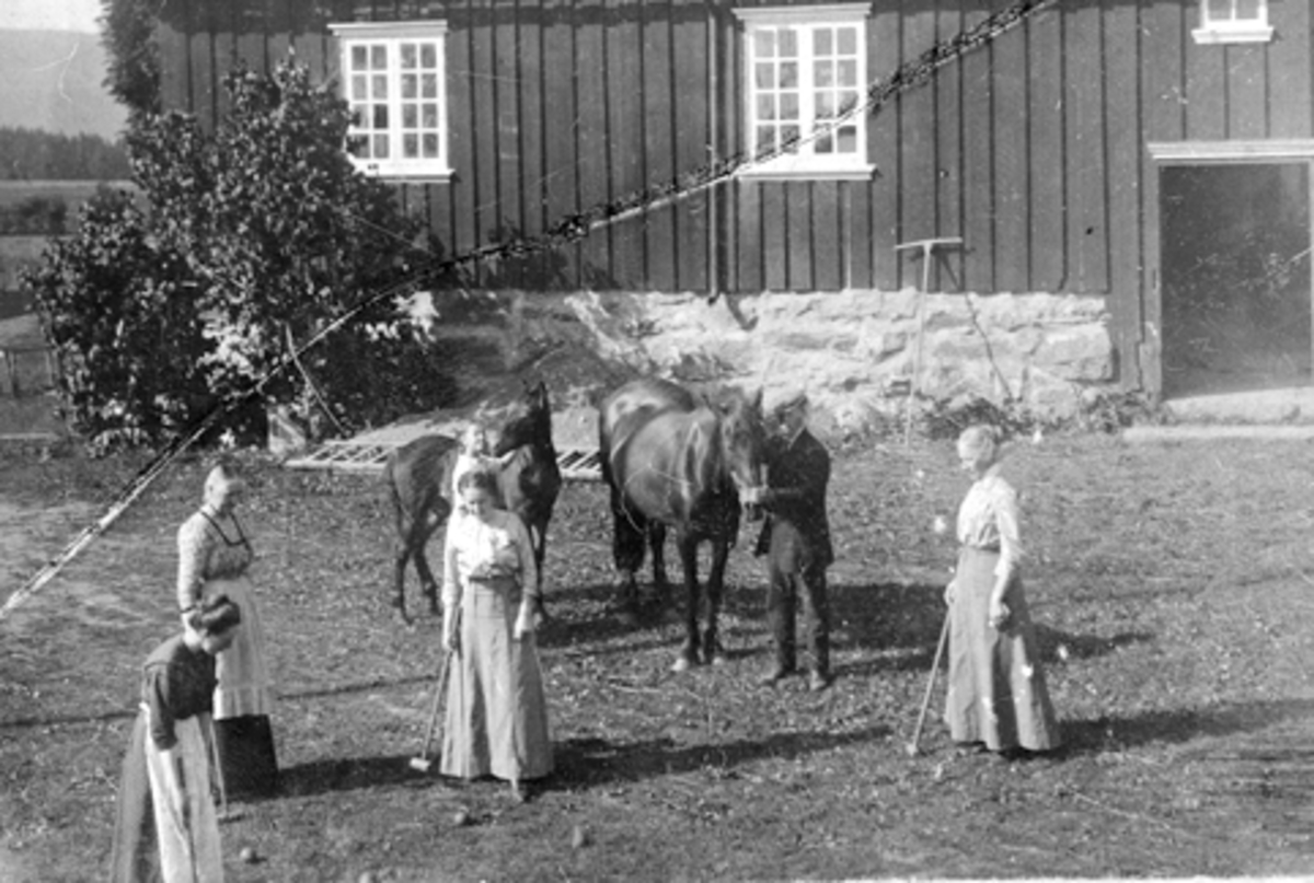 Gruppe kvinner spiller krokket foran drengestua på Mengshoel gard. Mann med to hester på tunet.