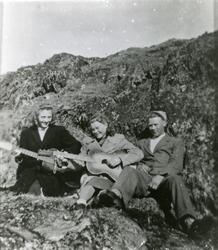 To kvinner og en mann sitter ved en bergknaus med gitarer