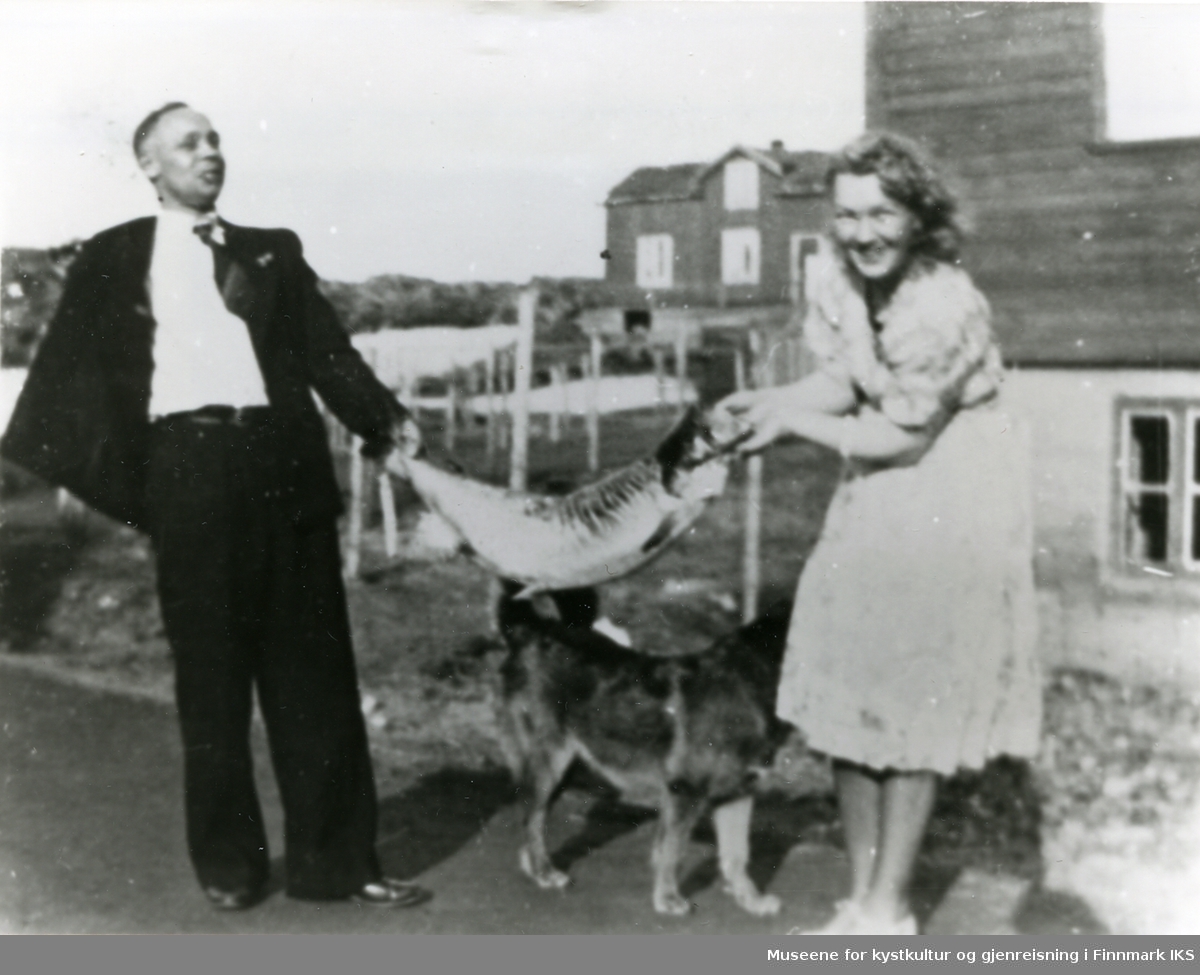 Dr. Vestby og frue står på en veg og holder en laks mellom seg, en hund og bebyggelse i bakgrunnen.