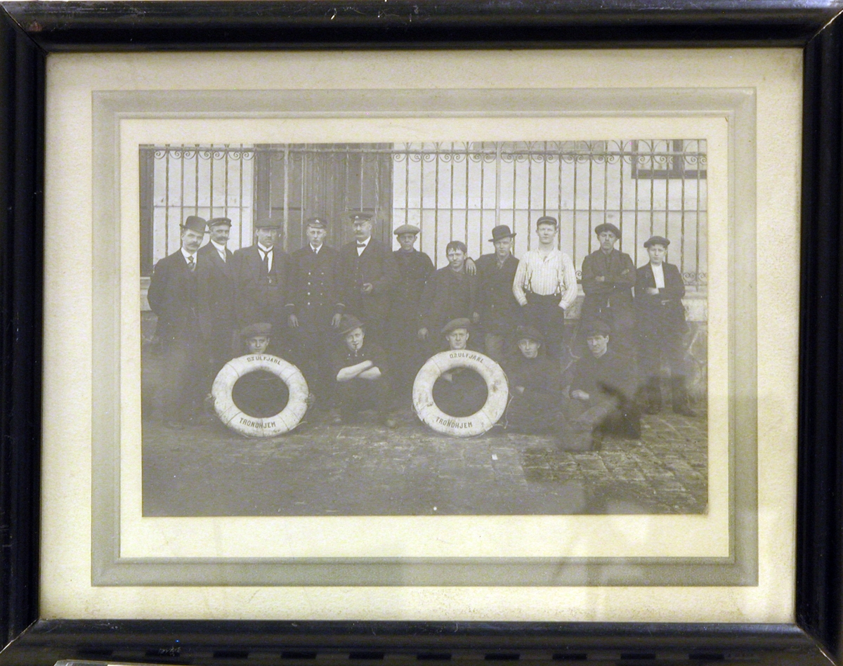 16 menn/gutter stående og sittende foran et murhus m/jerngjerde. To av mennene/guttene holder hver sin redningsbøye hvorpå det står skrevet: "D.S. ULF JARL  TRONDHJEM".