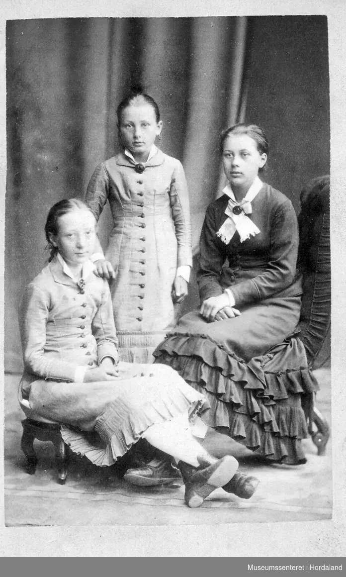 Tre jenter, ca. 8-15 år, i halvlange kjoler med kappeskjørt.