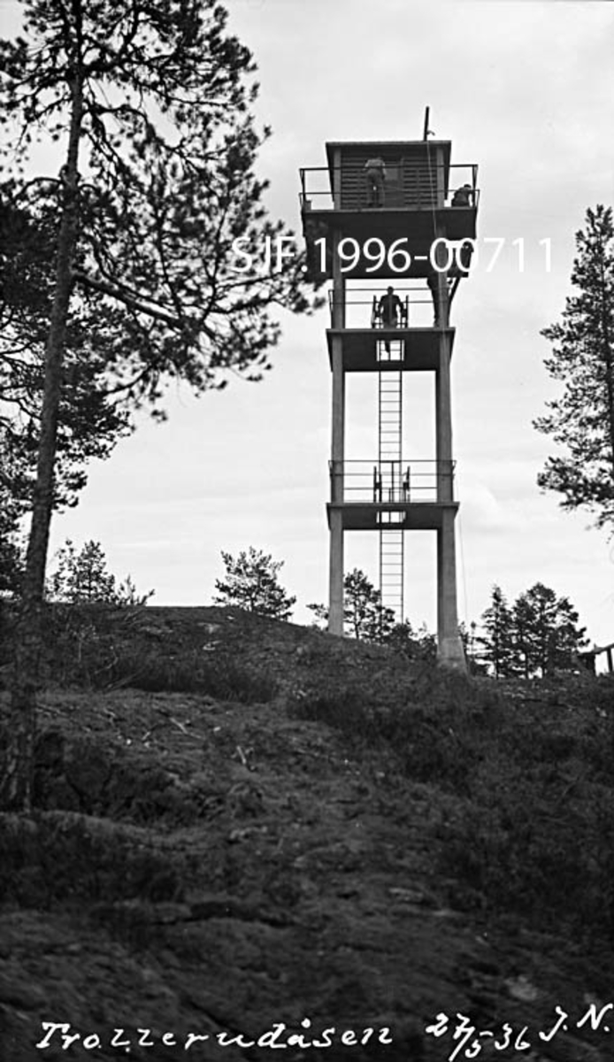 Skogbrannvaktstasjonen på Trollerudåsen i Spydeberg.  Brannvakttårnet på dette stedet ble reist i 1935, som en erstatning for eldre brannvaktposter på Kjerringa, Spydeberg varde og Bjørnåsen.  Trollerudås-tårnet er i hovedsak bygd av stål og betong, værbestandige materialer som skulle gi tårnet lang levetid.  Konstruksjonen består av fire loddrette betongsøyler med tre horisontale avsatser mellom.  Samtlige avsatser har stålrekkverk. På den øverste avsatsen er det bygd ei lita utkikkehytte av tre.  Atkomsten til hytta skjer ved hjelp av stiger som er ført gjennom åpninger i de ulike avsatsdekkene.  Dette fotografiet er tatt på forholdsvis kort hold, litt nedenfra, omtrent slik tårnet må ha fortont seg for besøkende.  En mann skimtes på "andre avsats", en annen ved rekkverket utenfor hytta på topplanet.  Omkring tårnet vokser det spredte furutrær. 