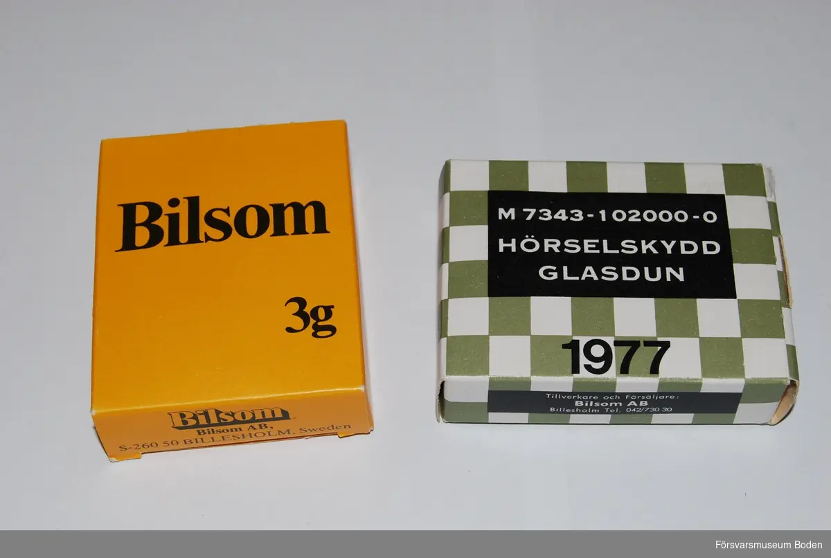 Två förpackningar med glasdun för öronproppar. Den ena är en civil variant med gul ask, och den andra försvarets ask m/1960 med materielnummer M7343-102000-0.