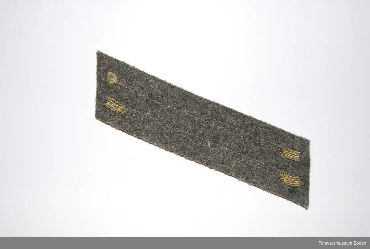Två band av bronsgalon på bakgrund av gråbrungrönt kommisskläde. Användes till vapenrock m/1923 och syddes fast på vänster överärm 150 mm från axelsömmen.