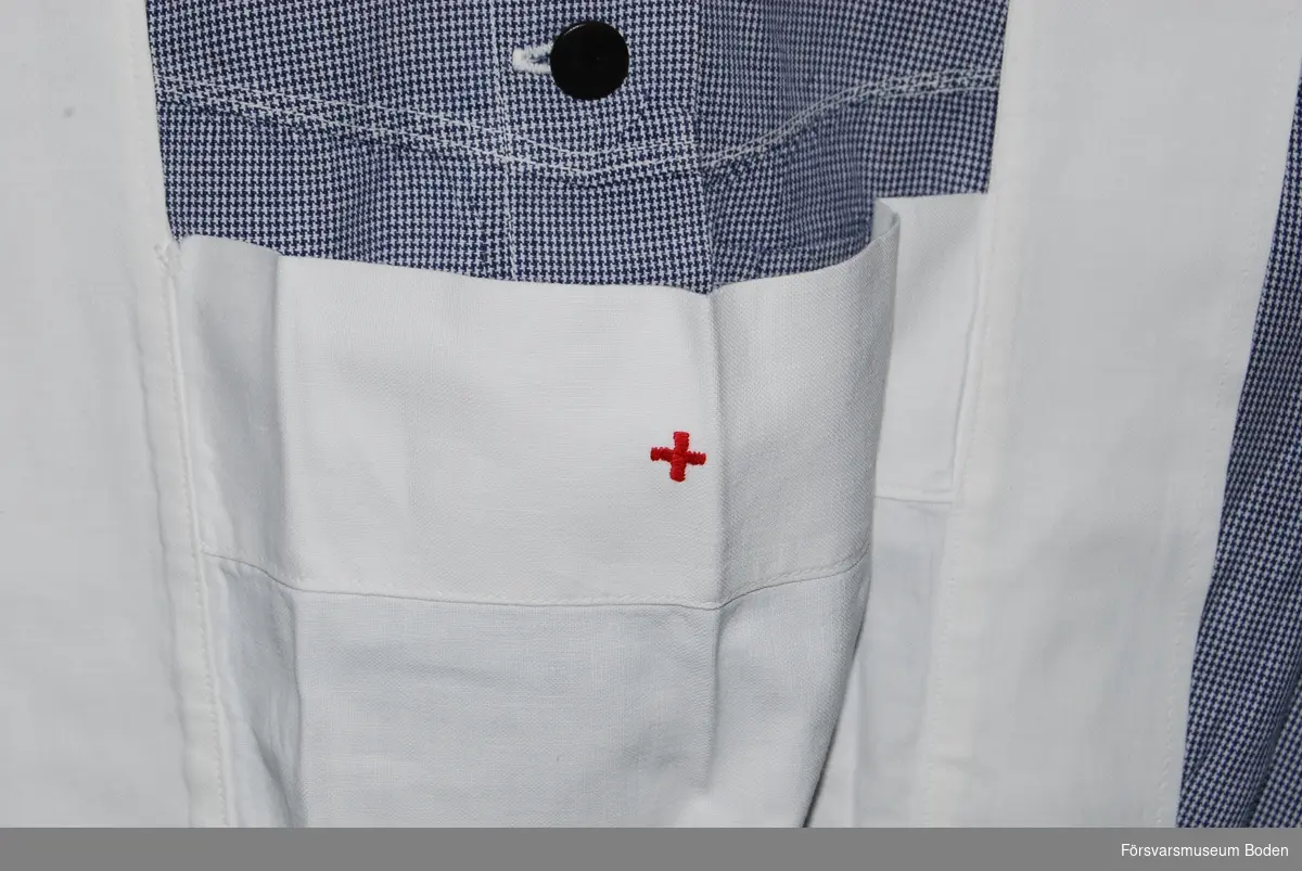Vitt bomullstyg med broderat rött kors i litet format framtill. Utanpåliggande ficka på höger sida.