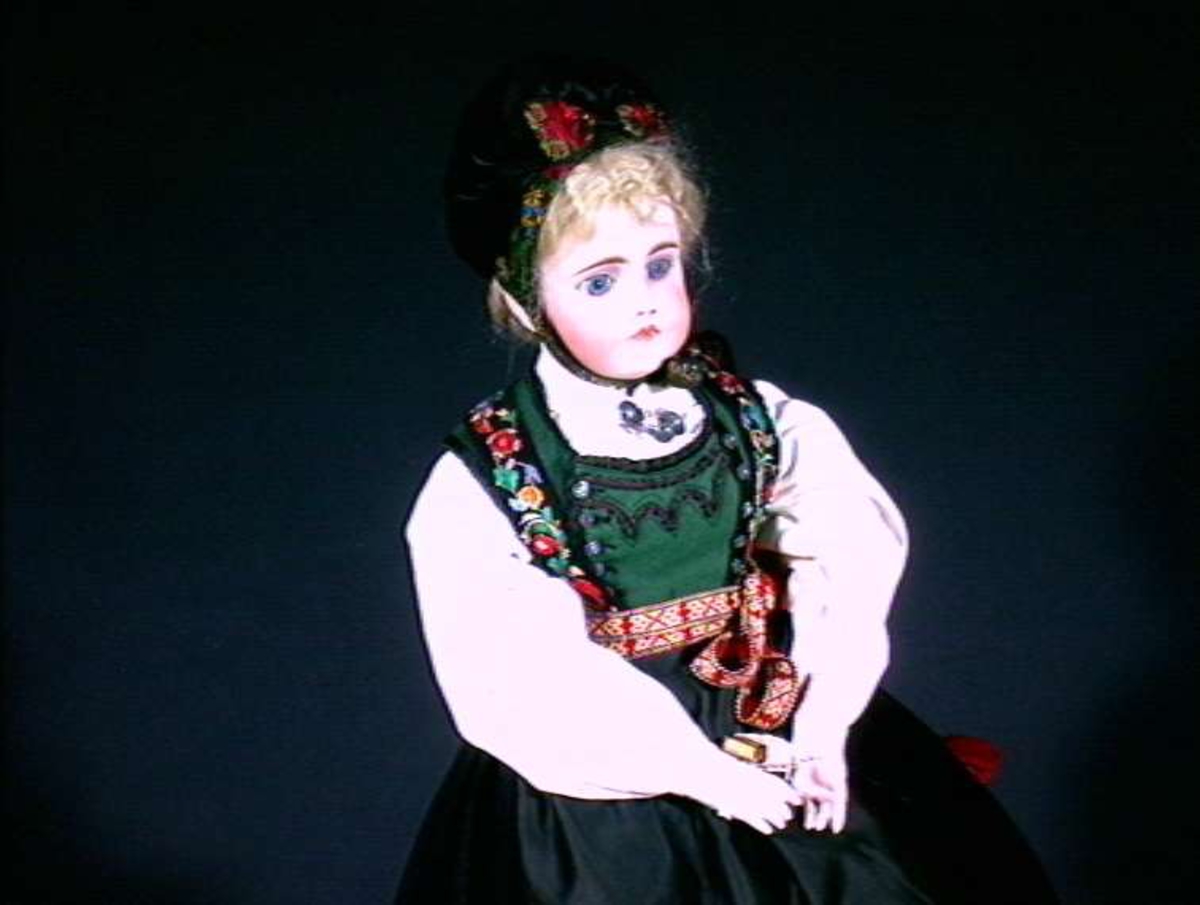 Bunadsdukke med porselen hode og hender, blondt hår.
Dukken er kledd i stakk med liv, forkle og lue med pannelin (B-Q), Telemark.