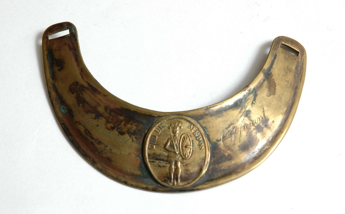 Sigdformet ringkrave i messing med oval medaljong i sentrum.
Medaljonen har innskrift og viser en gudinnefigur med sverd og skjold.